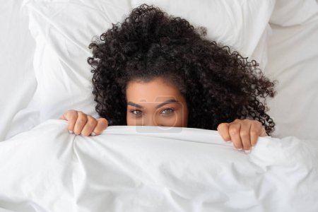 Eine Frau lugt unter einer Decke hervor, ihre Augen sind neugierig. Der kuschelige Stoff bedeckt teilweise ihr Gesicht, während sie sich diskret umsieht.
