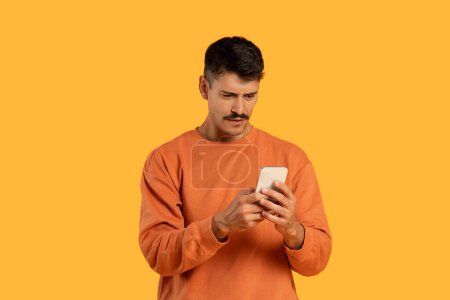 Foto de Un hombre con una camisa naranja está de pie mientras está enfocado en la pantalla de su teléfono celular, conectado con el dispositivo, posiblemente desplazándose o leyendo. El fondo es neutral, enfatizando su actividad. - Imagen libre de derechos