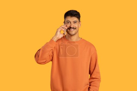 A man wearing an orange sweater is engrossed in a phone conversation. He holds a cell phone to his ear and gestures animatedly as he talks.