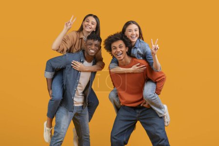 Photo for A joyful multiethnic group of friends piggybacking, showing peace signs, and laughing, isolated on an orange background portraying happiness and team spirit - Royalty Free Image