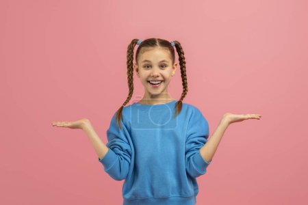 Téléchargez les photos : Une adolescente souriante dans un pull bleu les mains levées, heureuse et ludique, isolée sur un fond rose - en image libre de droit