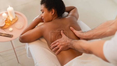 Téléchargez les photos : Une femme afro-américaine profite d'un massage relaxant du dos par un thérapeute professionnel dans un environnement spa serein comprenant des bougies et une esthétique confortable - en image libre de droit