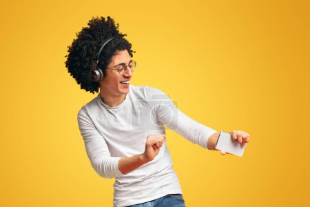 Photo for Active carefree black guy in wireless headphones enjoying music online on smartphone and dancing, orange background - Royalty Free Image