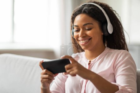 A cozy living room setting captures a serene moment of a young African American woman immersed in music, using her phone and headphones while seated comfortably on a couch, emanating a peaceful vibe