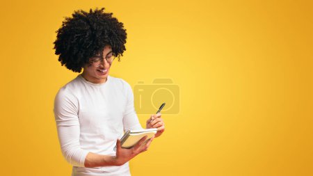 Foto de Lindo afroamericano millennial chico tomando notas, escritura lista de verificación en cuaderno, fondo naranja - Imagen libre de derechos