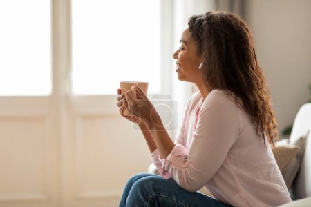 A relaxed African American woman sits on a soft couch, wearing earphones and listening intently to music, drinking coffee, exuding a calm and peaceful demeanor in her home setting