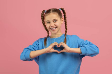 Teenager showcasing a heart gesture, smiling warmly, isolated on pink, embodying love and positive feelings in a youngsters portrait