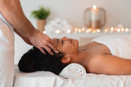 Eine afroamerikanische Dame erhält eine Kopfmassage in einem ruhigen Wellnessbereich mit Kerzen, Seitenansicht. Körperpflegekonzept