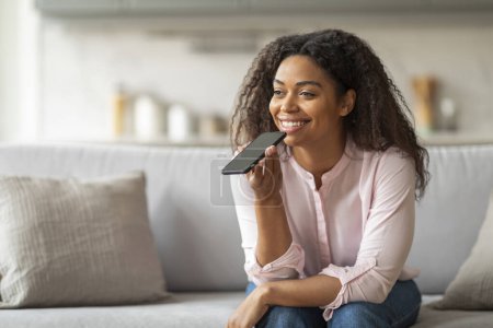 Eine fröhliche junge Afroamerikanerin bedient ihr Smartphone bequem per Sprachbefehl, während sie es sich zu Hause auf dem Sofa gemütlich macht