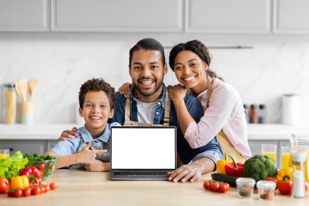 Téléchargez les photos : Famille noire aimante souriante montrant ordinateur portable avec écran blanc vierge, cuisine ensemble à la cuisine, espace de copie de maquette - en image libre de droit