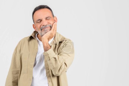 Photo for A senior man sits with his hand placed on his face, suffering from toothache on white background, copy space - Royalty Free Image