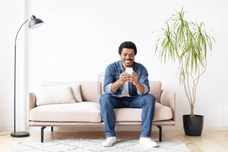 Téléchargez les photos : Joyeux homme afro-américain à la maison texter sur son smartphone, exprimant la communication et le bonheur - en image libre de droit