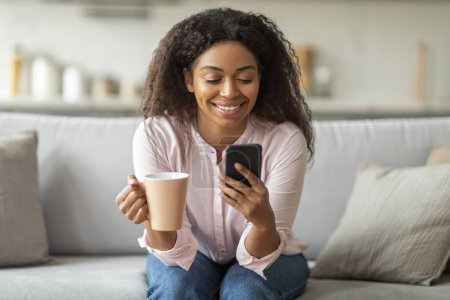 Une scène de l'après-midi détendue où la femme afro-américaine profite de son temps libre en s'engageant avec son appareil mobile, assise confortablement sur son canapé du salon, buvant du café