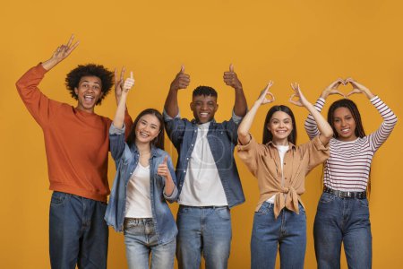 Téléchargez les photos : Cinq jeunes adultes multiethniques joyeux montrant divers gestes positifs de la main sur un fond orange vif - en image libre de droit