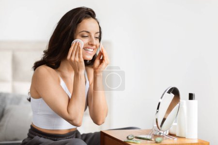 Téléchargez les photos : Jeune femme souriante en tenue décontractée utilisant un coton pour nettoyer son visage, assise à une coiffeuse avec des cosmétiques - en image libre de droit