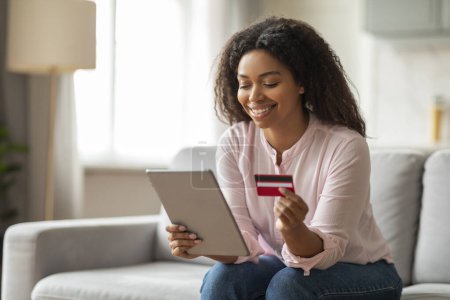 Téléchargez les photos : Joyeux jeune femme afro-américaine avec carte de crédit en utilisant une tablette numérique à la maison, assis sur le canapé dans le salon confortable intérieur - en image libre de droit
