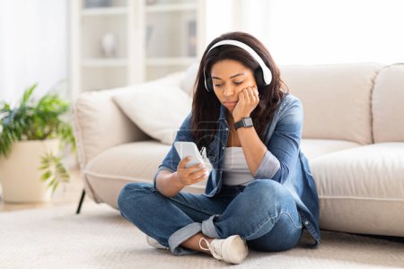 Foto de Mujer afroamericana sentada en el suelo en interiores, usando un teléfono celular y auriculares, buscando contenido interesante para ver - Imagen libre de derechos