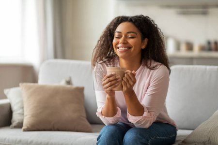 Photo for Positive young black woman sitting alone on couch at home, drinking coffee with closed eye and smiling. Millennials lifestyle - Royalty Free Image