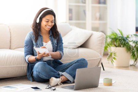 Photo for African American woman is sitting on the floor with crossed legs, focused on her laptop screen, have video lesson or attending webinar - Royalty Free Image