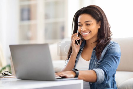 Photo for Busy African American woman sits at a desk, multitasking by talking on a cell phone while typing on her laptop. She appears focused and engaged in her work, balancing communication and productivity. - Royalty Free Image