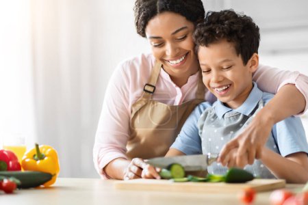 Téléchargez les photos : Moment chaud comme une mère afro-américaine enseigne doucement à son fils comment couper les légumes dans la cuisine, la promotion de la famille et l'apprentissage - en image libre de droit