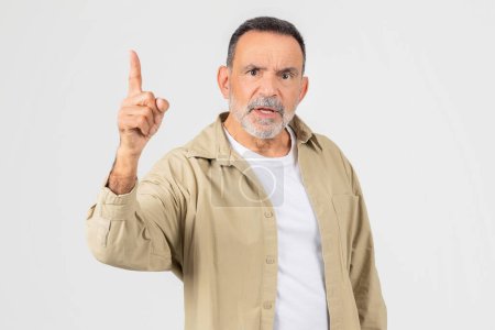 A mature gentleman with a beard stands against a plain background, looking directly at the viewer while assertively raising his index finger