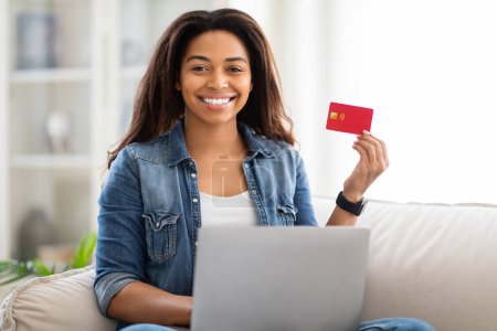 African American woman is seated on a couch, holding a credit card and a laptop in her hands, engaging in online shopping or financial transactions