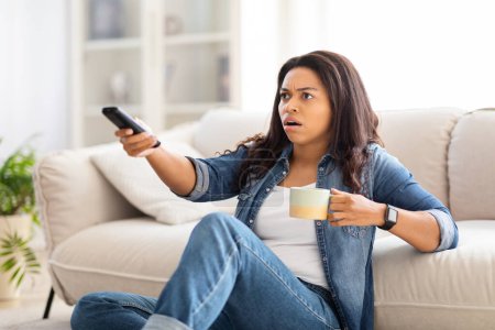 Photo for Shocked African American woman is seated on a couch, holding a remote control in her hand, possibly changing channels or adjusting the volume - Royalty Free Image