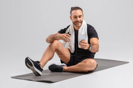 Téléchargez les photos : Un homme est assis sur un tapis de fitness noir, tenant une bouteille d'eau, reprenant son souffle après une séance d'entraînement. Porter un short de sport, un haut sans manches et des chaussures de sport, en utilisant un smartphone - en image libre de droit