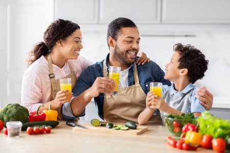 Foto de Alegre familia afroamericana padre madre e hijo bebiendo jugo de naranja fresco mientras cocinan comida saludable juntos en la cocina - Imagen libre de derechos