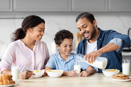 Téléchargez les photos : Une famille afro-américaine partage un moment de lumière tandis que le père verse du lait dans un verre, incarnant la chaleur de la vie familiale dans un cadre de cuisine - en image libre de droit
