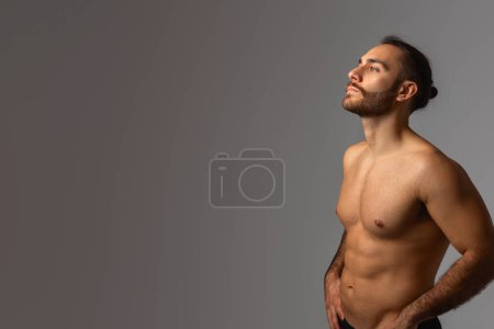 A male figure without a shirt, standing upright with a neutral expression against a plain gray backdrop. The mans physique and lack of clothing are the focal points of the image, copy space