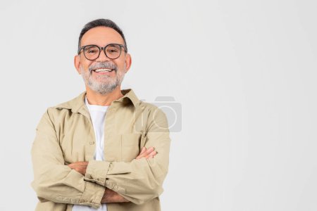 Photo for A senior man wearing glasses is standing upright with his arms crossed over his chest. He appears confident and assertive, with a serious expression on his face, copy space - Royalty Free Image
