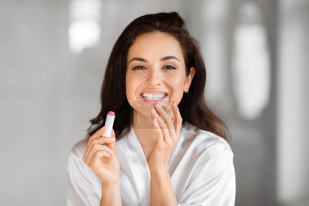 Foto de Una mujer radiante en un albornoz blanco sonríe mientras aplica lápiz labial, personificando rituales milenarios de cuidado de la piel del baño - Imagen libre de derechos