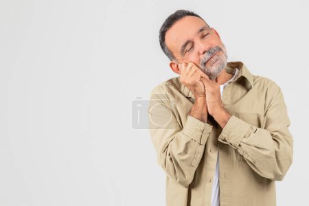 Photo for A senior man is standing with his hands pressed together against his face, feeling sleepy on white background, copy space - Royalty Free Image