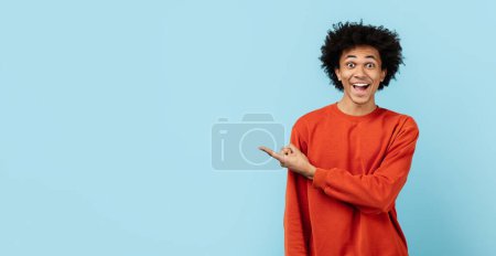 A lively black guy with a wide smile, excitedly pointing at copy space, wearing an orange sweater against a blue isolated background