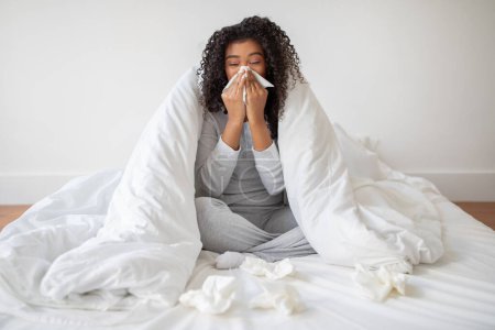 Photo for A young woman wrapped in a white duvet appears to be suffering from a cold or flu, sitting on a bed with tissues scattered around her, as she blows her nose - Royalty Free Image