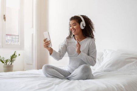 Eine fröhliche junge hispanische Frau sitzt im Schneidersitz auf einem weißen Bett, trägt gemütliche Freizeitkleidung und Kopfhörer und ist mit einem Videoanruf beschäftigt