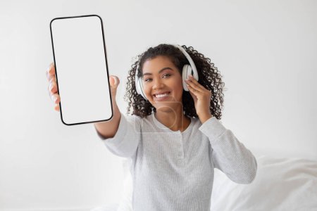Photo for A cheerful young woman with curly hair is holding a smartphone with a blank screen towards the camera, while simultaneously adjusting her white headphones with her other hand - Royalty Free Image