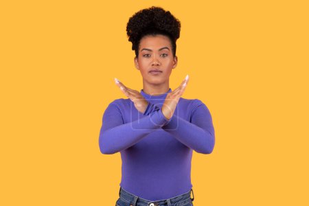 Hispanic young woman stands against a vivid yellow backdrop, crossing her arms in front of her to form an X, signaling stop or rejection