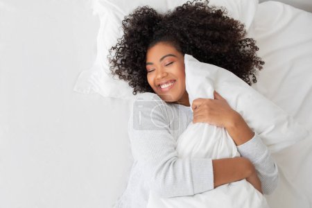 Eine Frau liegt in einem Bett, bedeckt mit einer weißen Decke, und umarmt ein Kissen. Der Raum ist sanft beleuchtet, was die friedliche Atmosphäre der Szene unterstreicht