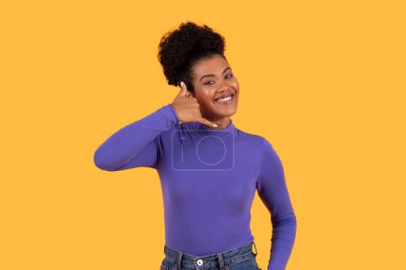 Foto de Hispanic woman wearing a purple shirt showing call me gesture with a focused expression. She appears to be standing on yellow background. - Imagen libre de derechos