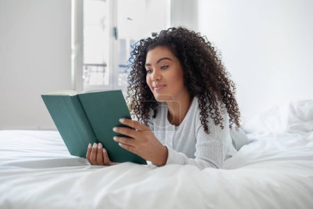 Photo for Hispanic woman is reclining on a bed with a book in her hands, absorbed in reading. The room is cozy and well-lit, creating a comfortable reading environment. - Royalty Free Image