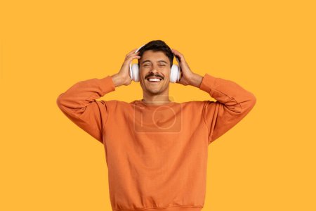 Foto de Un hombre con una camisa naranja está escuchando los auriculares, centrándose intensamente en la música que se reproduce. Su expresión es contemplativa mientras disfruta del sonido. - Imagen libre de derechos