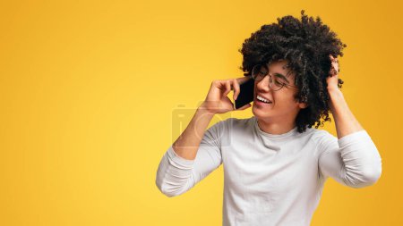 Téléchargez les photos : Timide homme afro-américain parlant sur smartphone, souriant et touchant ses cheveux, fond orange - en image libre de droit
