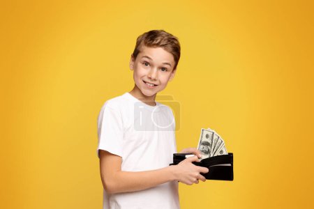 Rich teenager. Happy little boy holding wallet with lots of dollars, orange studio background