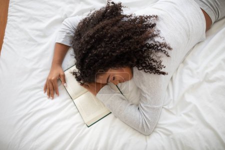 A young Hispanic woman has fallen asleep while reading a book in a comfortably bright bedroom. She is lying on her stomach on a white bed with the book still open under her arms