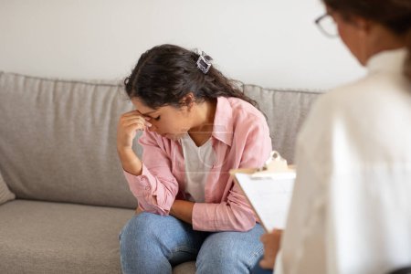 Photo for A young woman sits on a couch, her body language expressing distress as she covers her face with her hand. In the foreground, a therapist, indicated by the presence of a clipboard, offers a support - Royalty Free Image