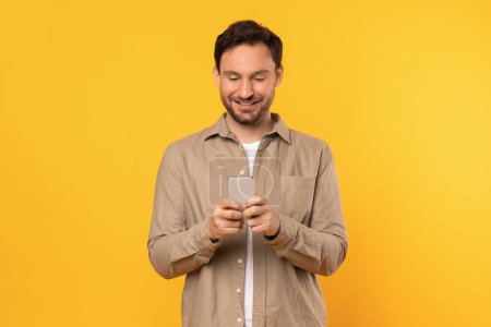 A man is standing in front of a vibrant yellow wall, holding a cell phone in his hand. He appears to be focused on the screen, possibly typing a message or browsing