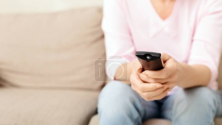 Lady est assise sur un confortable canapé beige avec les mains enroulées autour d'une télécommande de télévision, apparemment prête à changer de chaîne ou à ajuster le volume tout en profitant de temps libre à la maison.